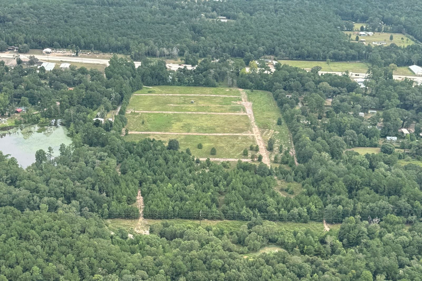 Aerial view of dense forest with a rectangular cleared tract in the middle, a pond at one corner, roads and scattered houses beyond