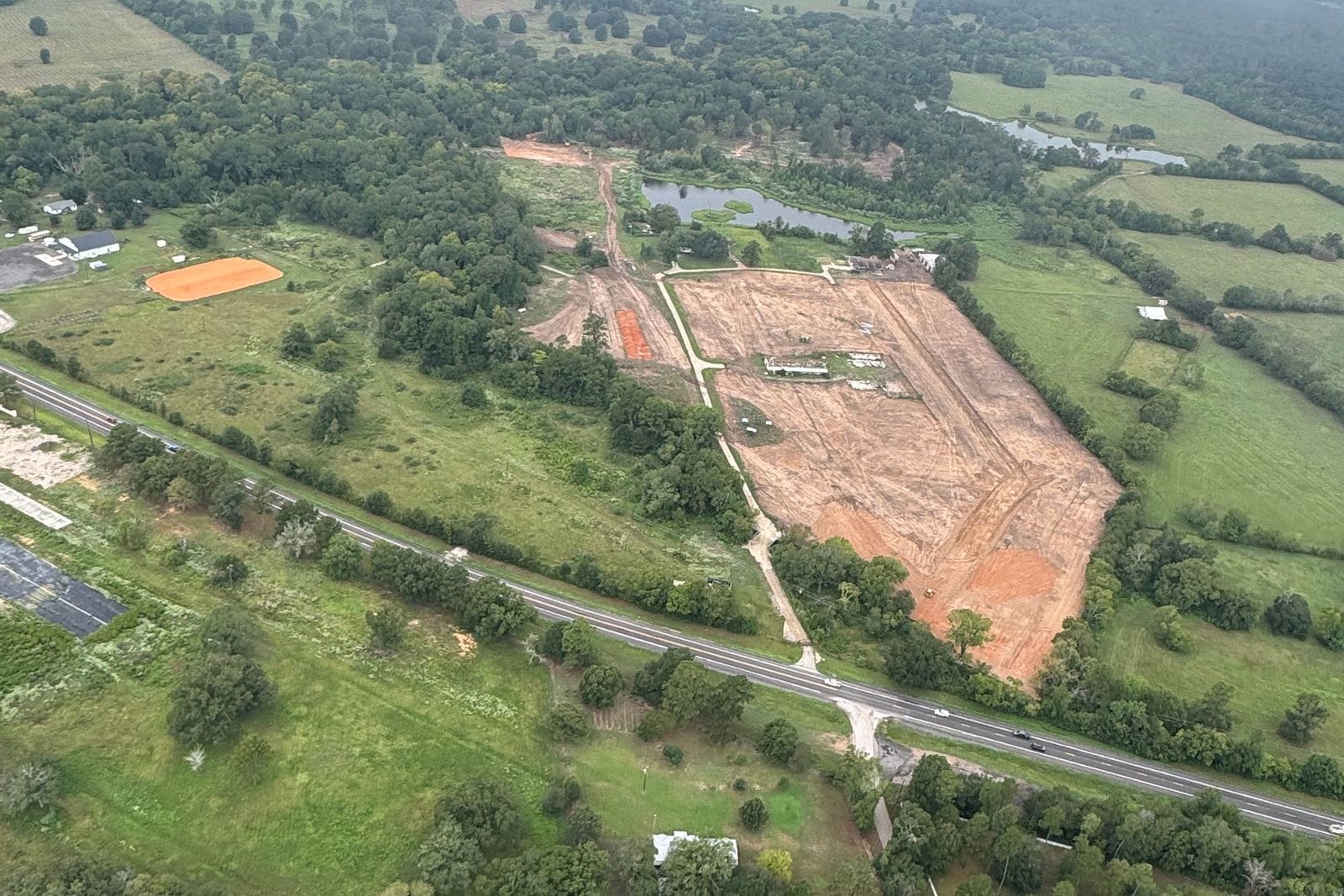 Aerial view of a tract being cleared and graded, bare orange earth beside a road, with pasture, houses and forest around it