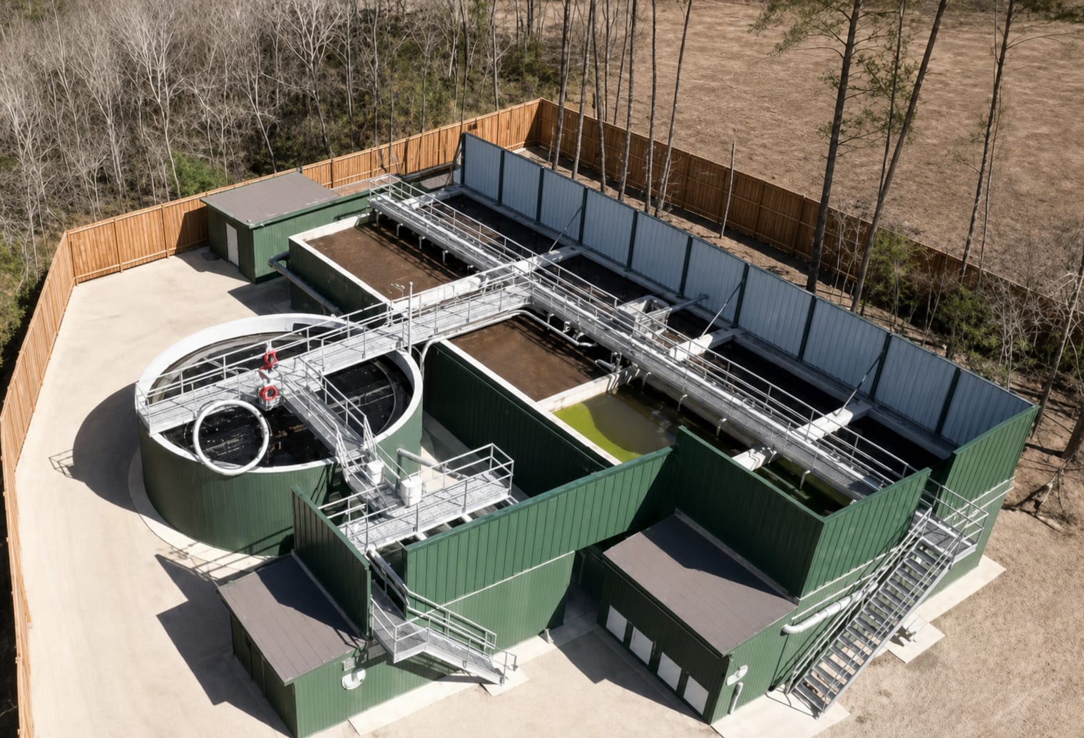 Aerial view of a wastewater treatment plant with green clad tanks, a circular clarifier, aeration basins and access walkways, surrounded by pine woodland